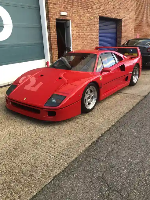 A red Ferrari F40 sports car is parked on a concrete surface in front of a brick building with garage doors. The car has a…