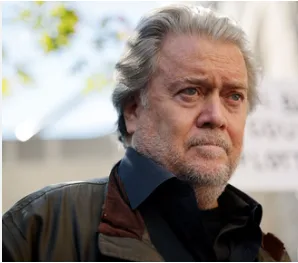 a close-up photo of a man with gray hair and a beard, wearing a dark jacket, looking off to the side with a serious…