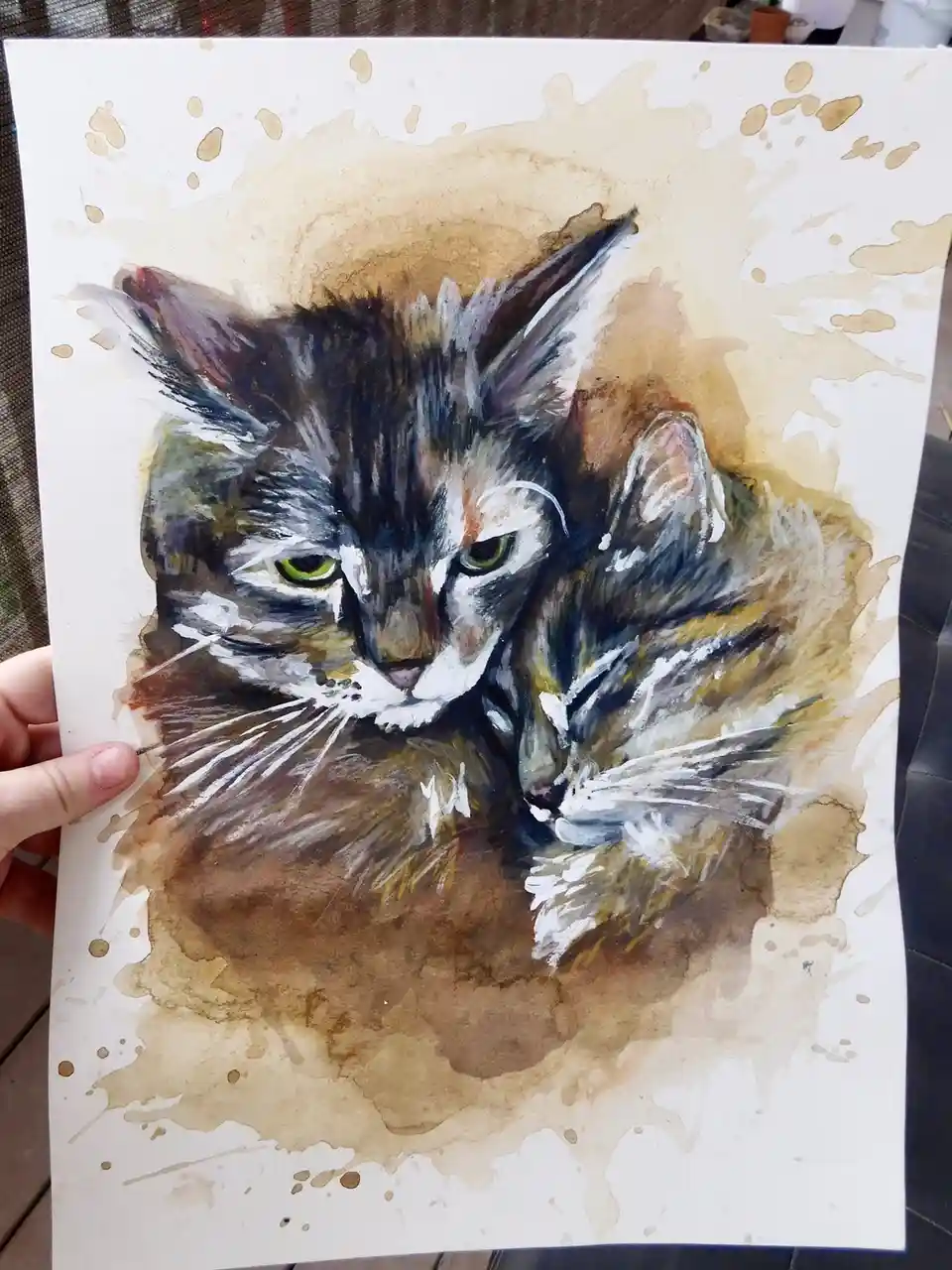 A hand holds up a watercolor painting of a cat with green eyes and a fluffy, multi-colored coat, set against a brown…