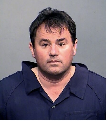 A mugshot of a man with dark hair and a neutral expression, wearing a dark blue collared shirt against a plain gray…