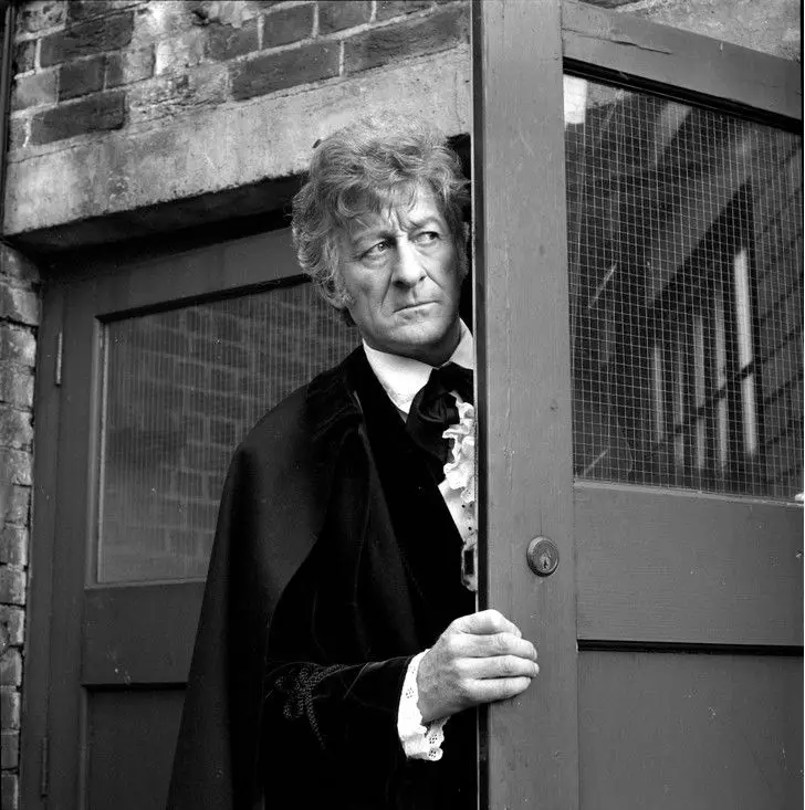 A black-and-white photo shows a man with curly hair in a dark velvet cape and ruffled shirt peeking from behind a wooden…