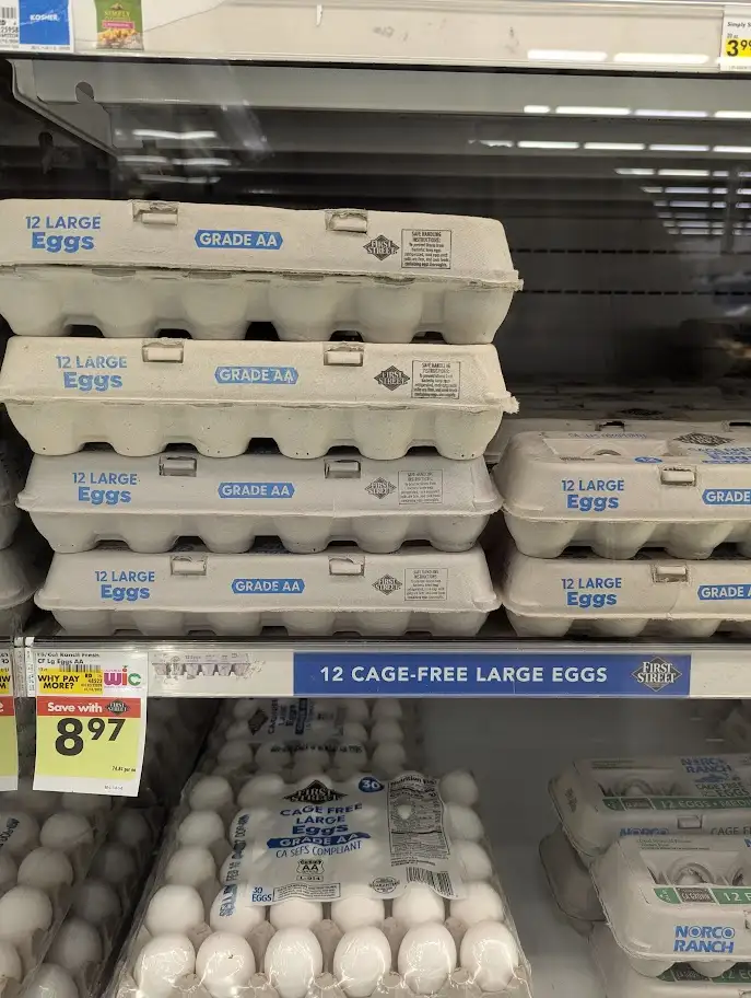 A grocery store egg aisle displays multiple stacks of 'Grade AA' egg cartons labeled '12 Large Eggs', with a blue shelf…