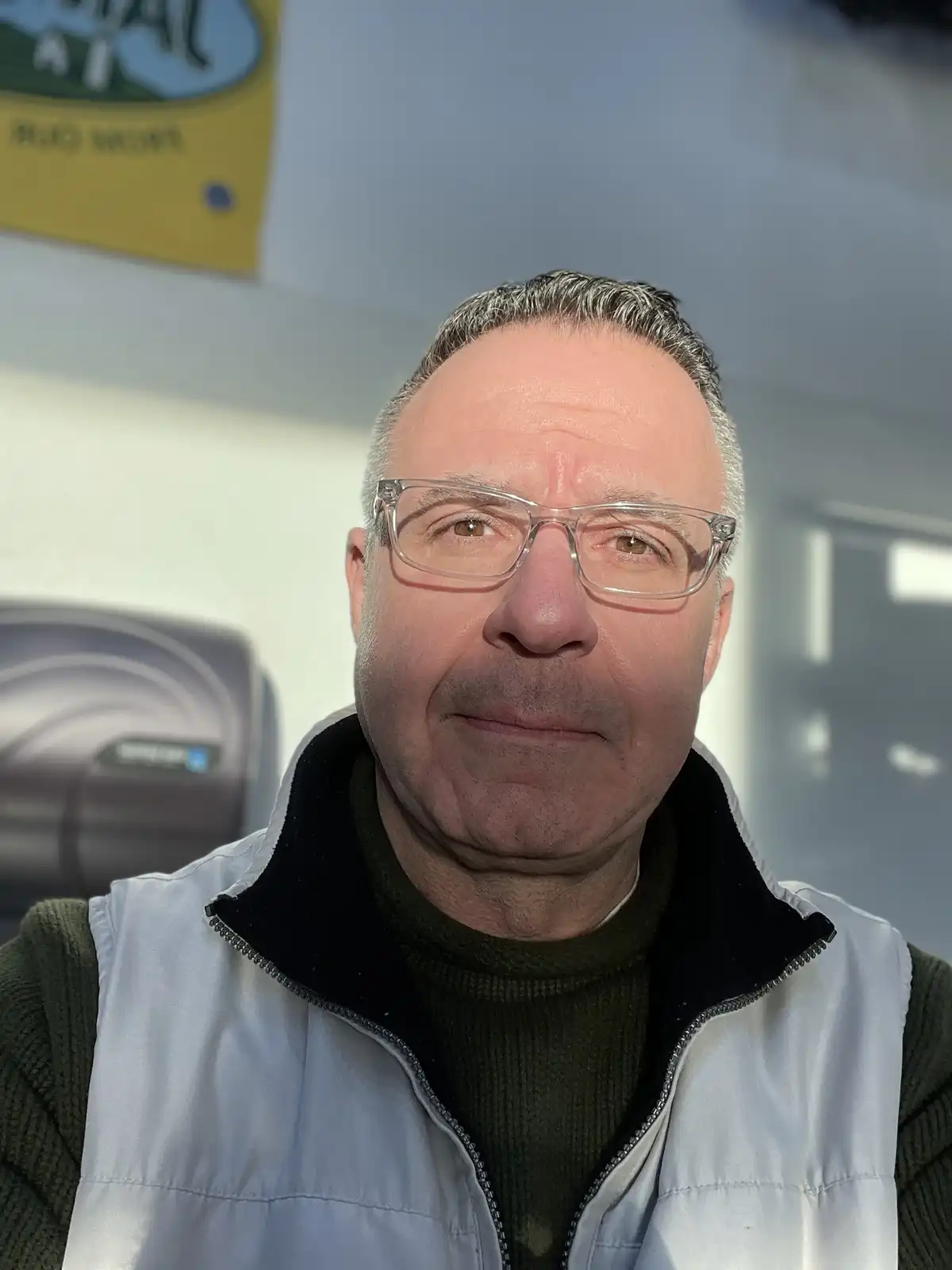 A middle-aged man with glasses and graying hair, wearing a light gray vest over a dark green turtleneck, stares directly at…