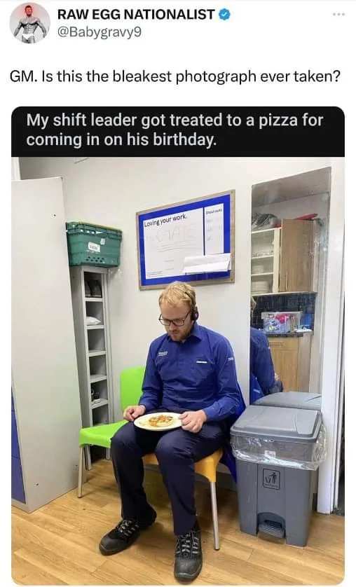 A man in a blue uniform and glasses sits on a green chair holding a plate with a single slice of pizza, looking dejected…