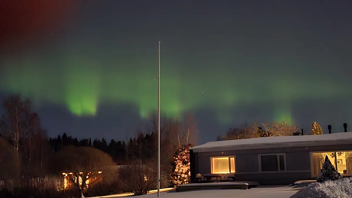 A snowy night scene shows a house with lit windows, bare trees, and a flagpole, all under a vibrant green aurora borealis…