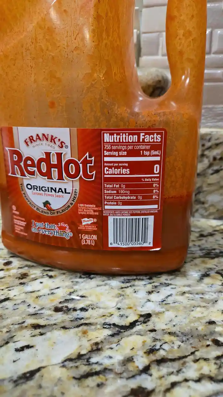 A close-up photo shows a large, 1-gallon plastic jug of Frank's RedHot Original Cayenne Pepper Sauce sitting on a granite…