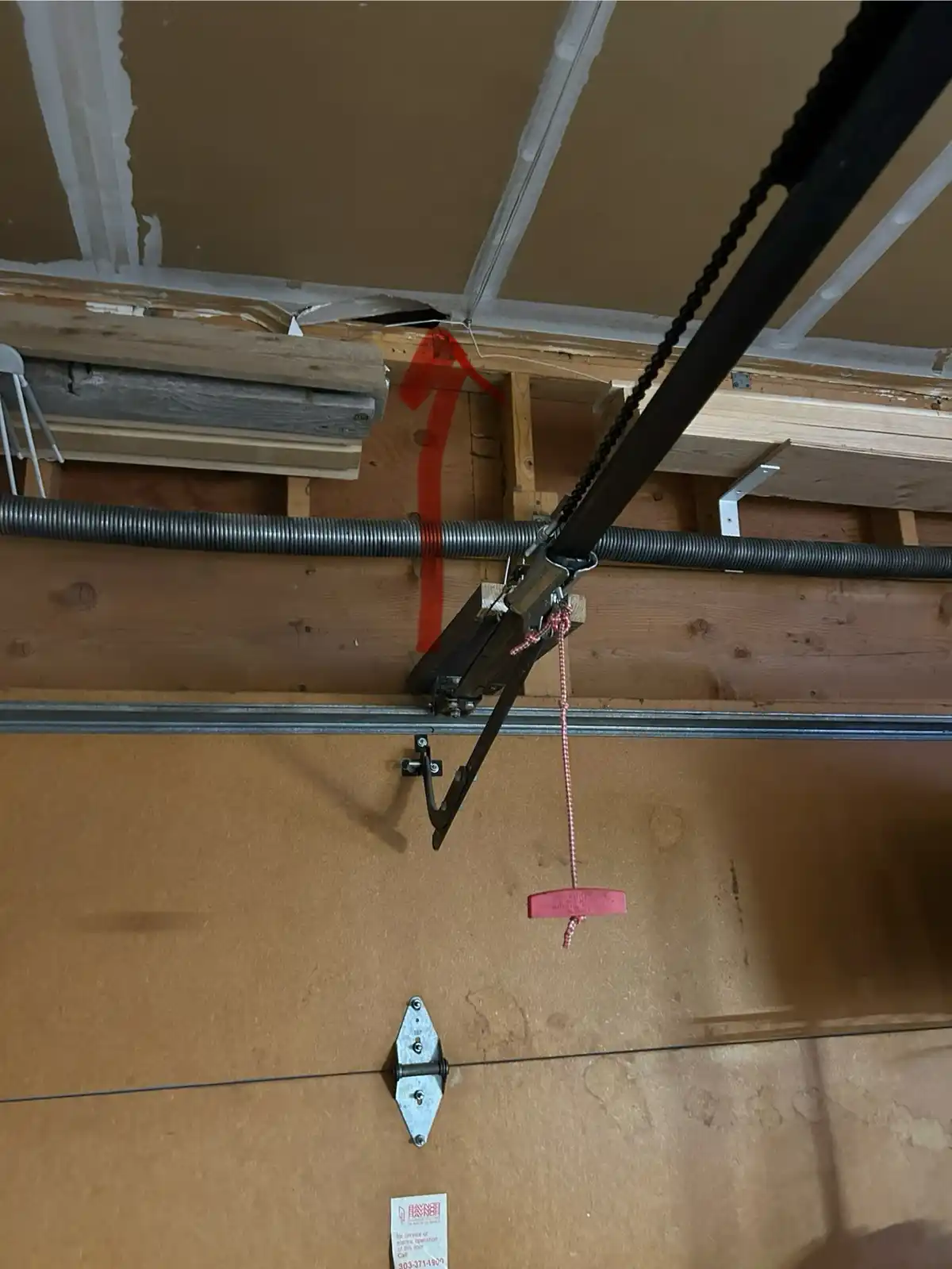 A close-up photo of a garage door opener mechanism with a red arrow drawn on the wooden ceiling joist pointing upward to a…