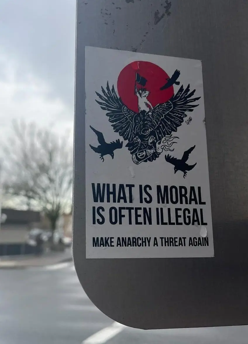 A sticker on a metal pole shows a black-and-white illustration of a winged, crowned figure holding a flag against a red sun…