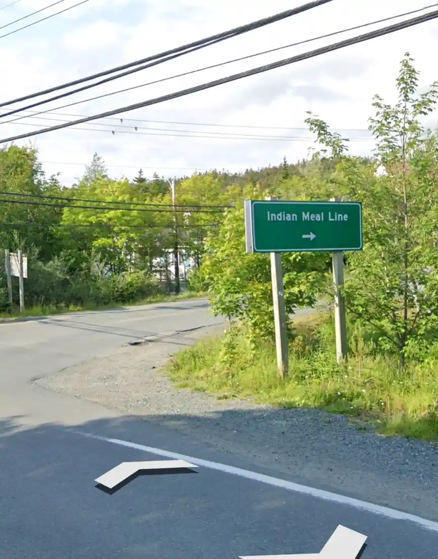 A green road sign in a rural area reads 'Indian Meal Line' with an arrow pointing right, while two white arrow graphics…
