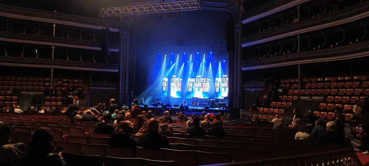 A wide shot of a large, ornate theater with multiple balconies and rows of seats, many of which are empty. The stage is lit…