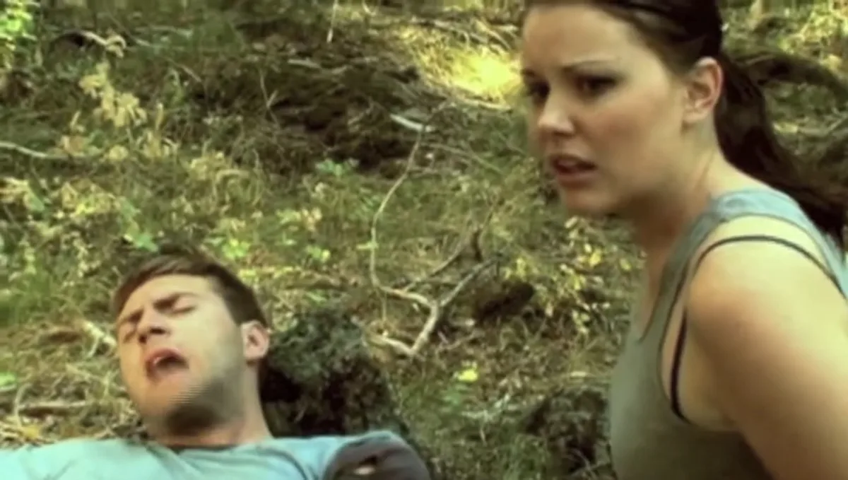 A man lies on the ground in a wooded area, mouth open as if yelling or in pain, while a woman in a tank top stands over him…