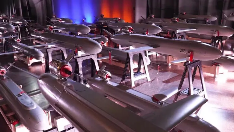 Rows of gray unmanned aerial vehicles (drones) are displayed on metal stands in a dimly lit room with colored stage…