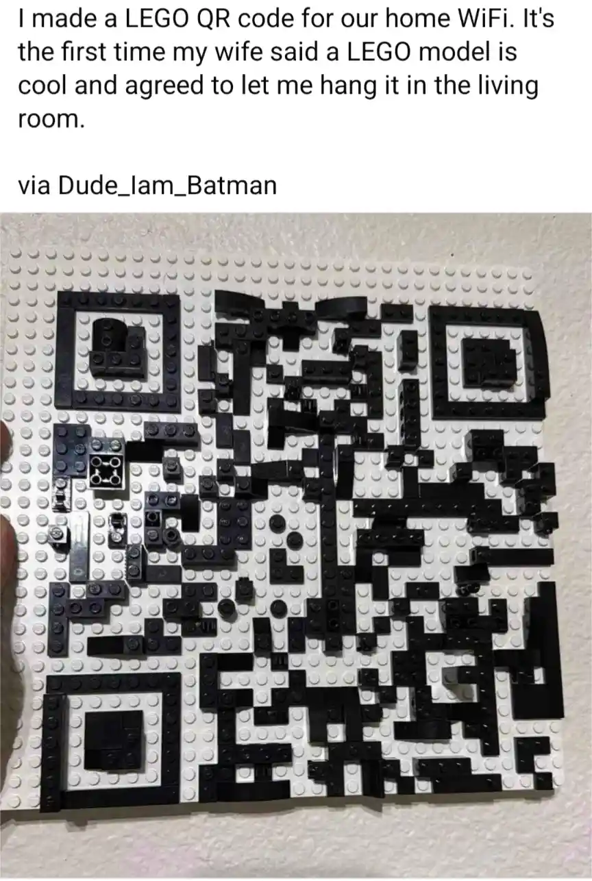 A hand holds a white LEGO baseplate with a black-and-white QR code constructed from LEGO bricks, mounted on a wall. The…