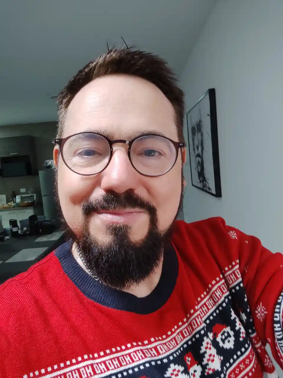 A man with glasses and a beard smiles at the camera while wearing a red ugly Christmas sweater with a Santa Claus pattern…