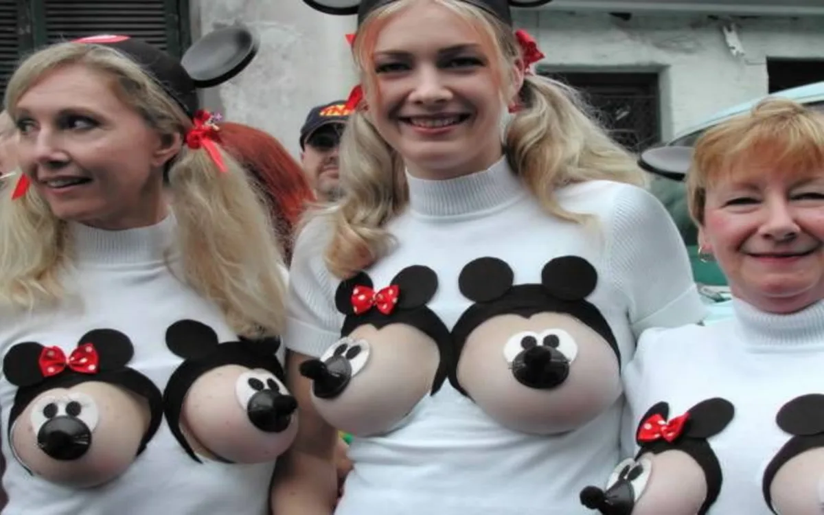 Three women wearing white turtleneck sweaters with Mickey and Minnie Mouse heads painted on their chests, using their…