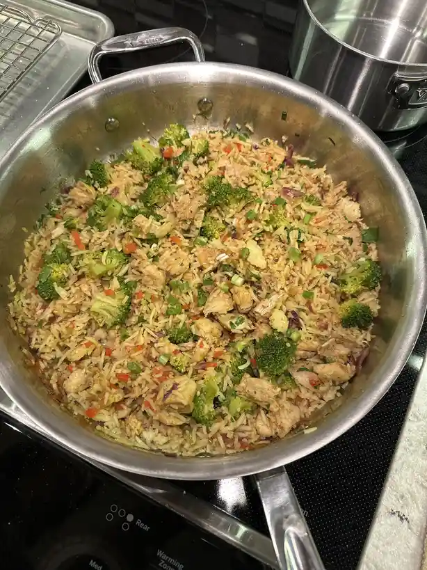 A stainless steel skillet on a stovetop contains a large serving of fried rice mixed with broccoli florets, chopped green…