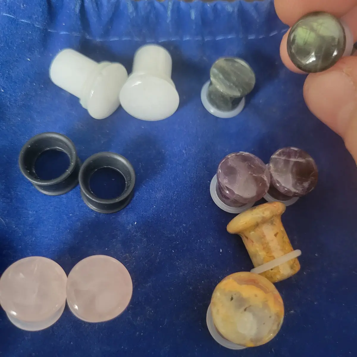 A hand holds a greenish translucent gemstone ear gauge over a blue fabric surface, surrounded by various other ear gauges…