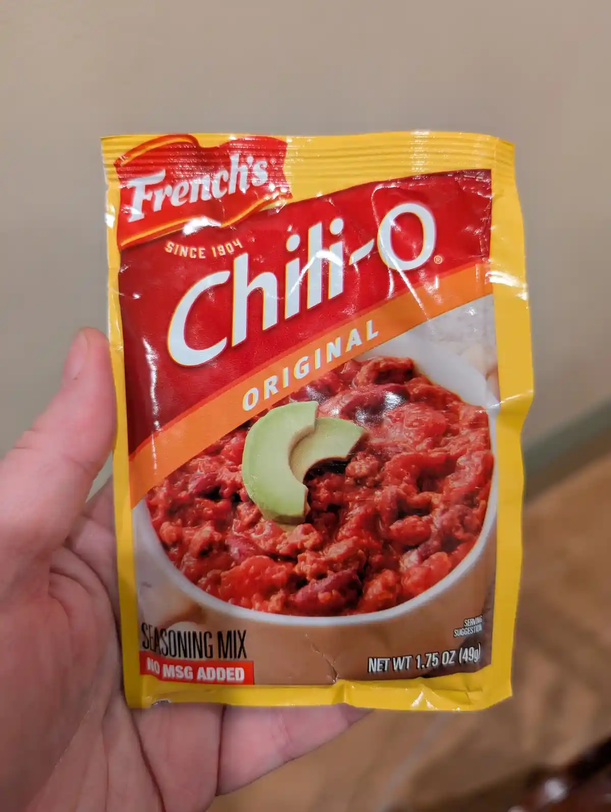 A hand holds a yellow and red packet of French's Chili-O Original seasoning mix, which features an image of chili topped…
