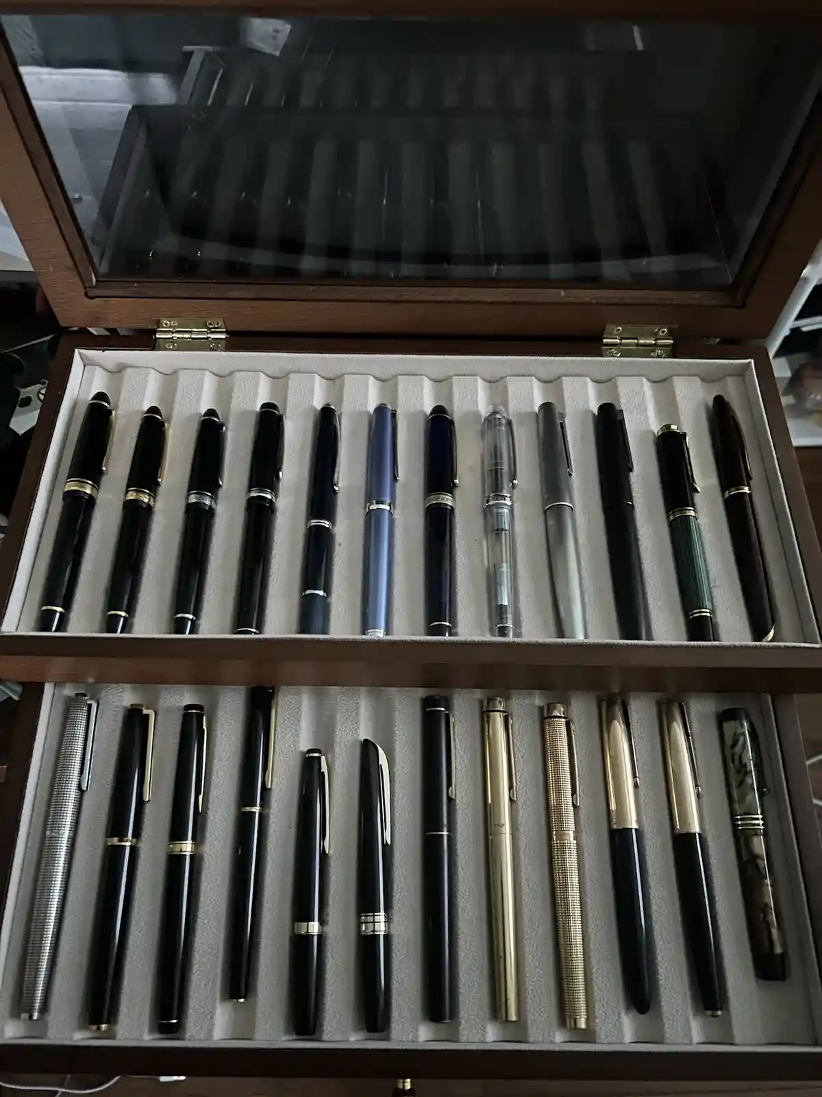 An open wooden display case with two rows of custom-fitted slots holds a large collection of pens, mostly black, silver, and…