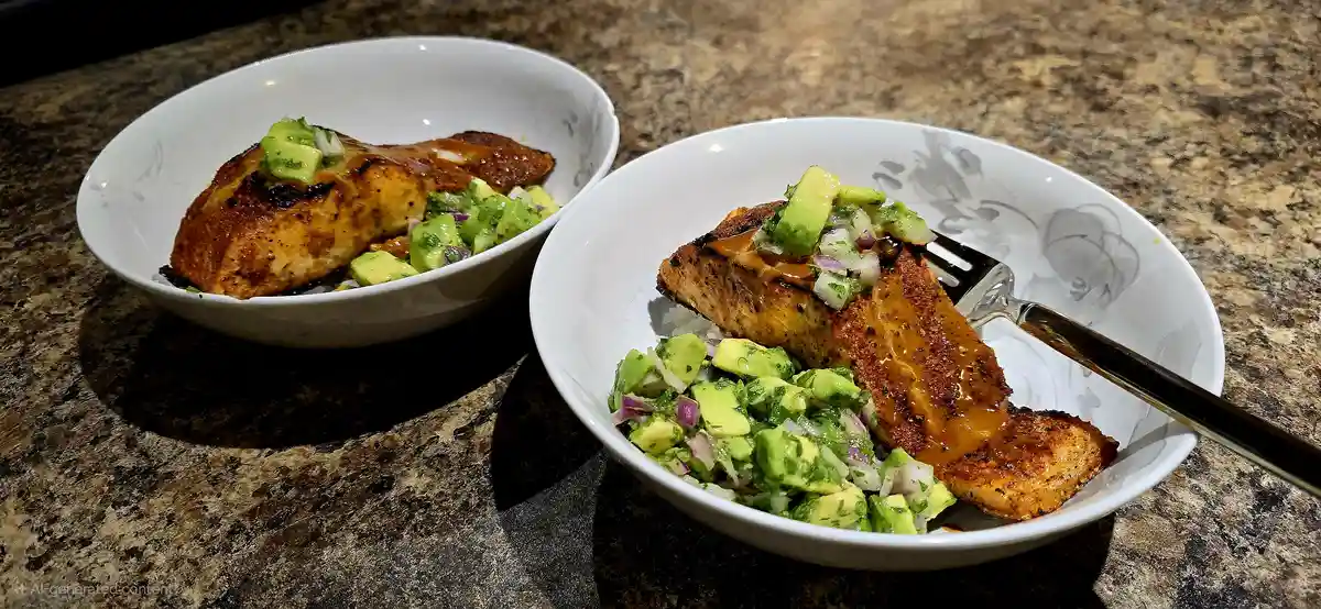 Two white bowls sit on a speckled granite countertop, each containing a grilled salmon fillet topped with avocado salsa and…