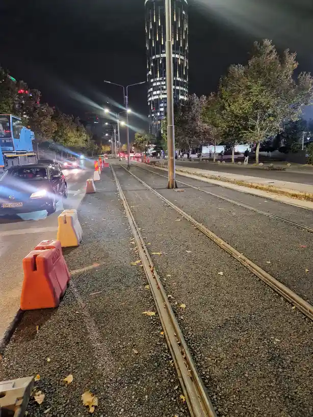 A nighttime street scene with tram tracks running down the center, flanked by orange and white traffic barriers. Cars with…