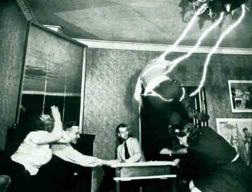 A black-and-white photo shows a man at a table being struck by a lightning bolt that appears to originate from above, while…