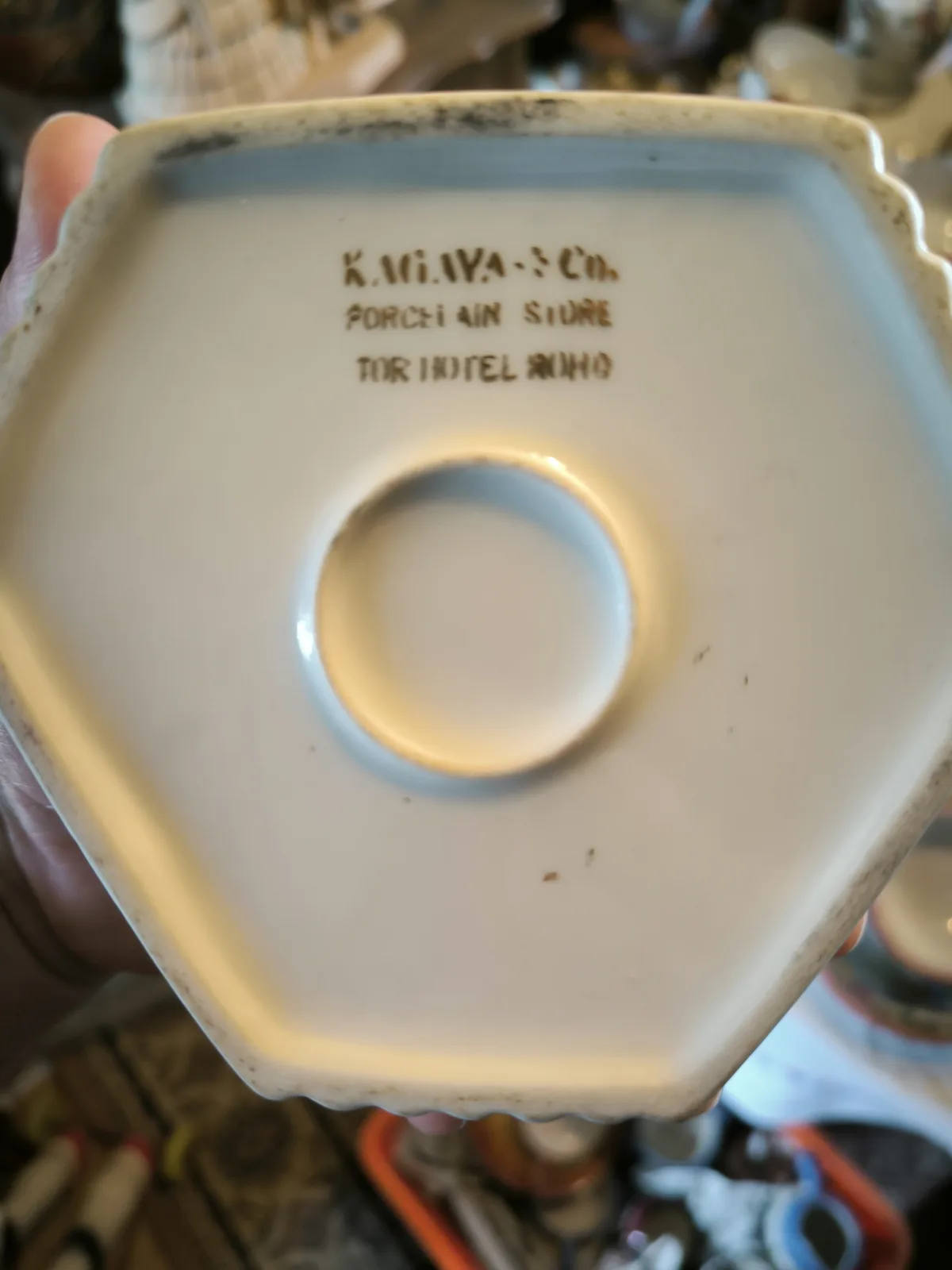 A close-up photo shows the bottom of a white ceramic item, likely a dish or vase, held in someone's hand. The base has a…