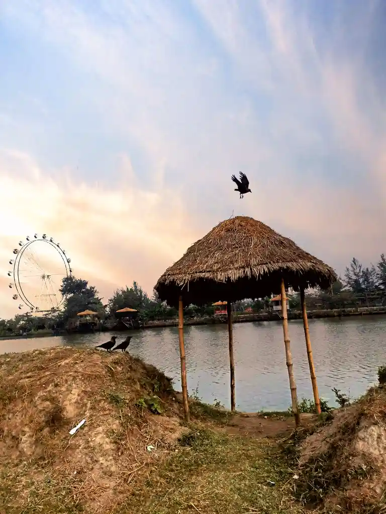 A black bird is captured mid-flight above a thatched-roof hut near a body of water, with two other crows perched on a nearby…
