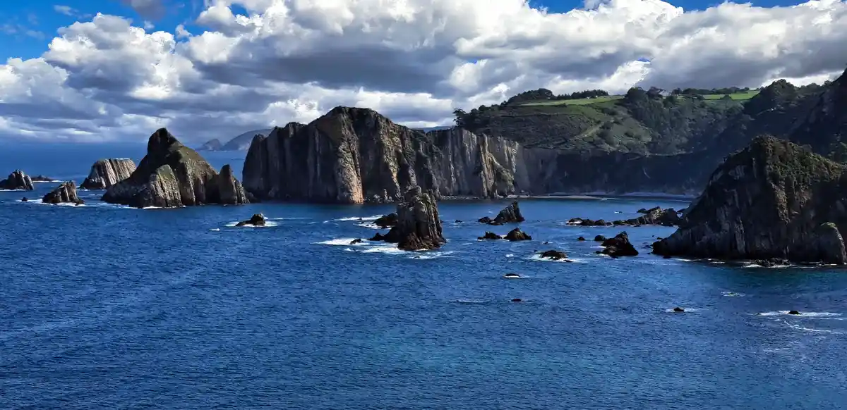 A scenic coastal landscape featuring deep blue ocean water with numerous dark, jagged rock formations and cliffs. The sky…