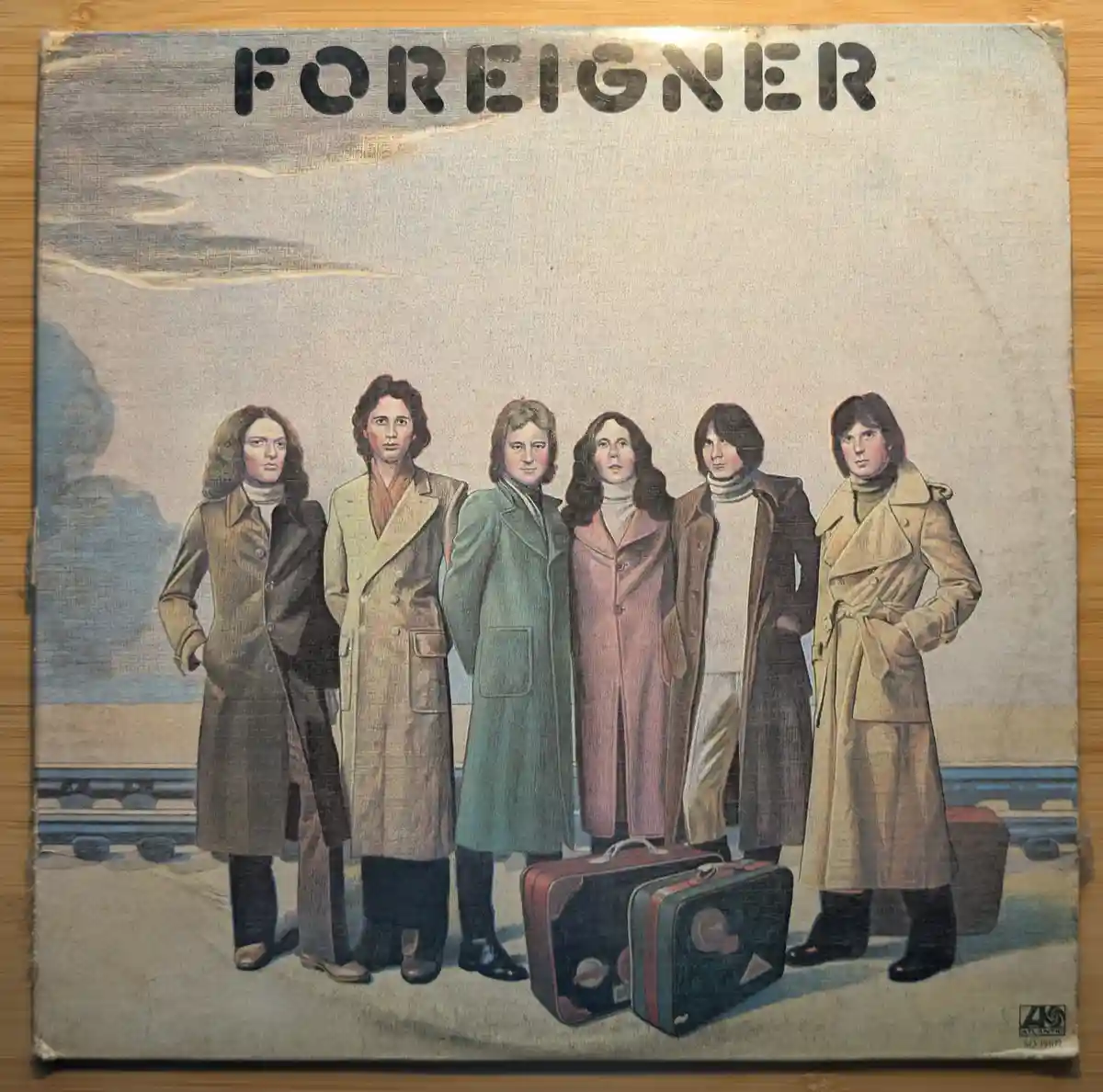 The image shows the album cover for the band Foreigner, featuring six band members in trench coats standing on a train…