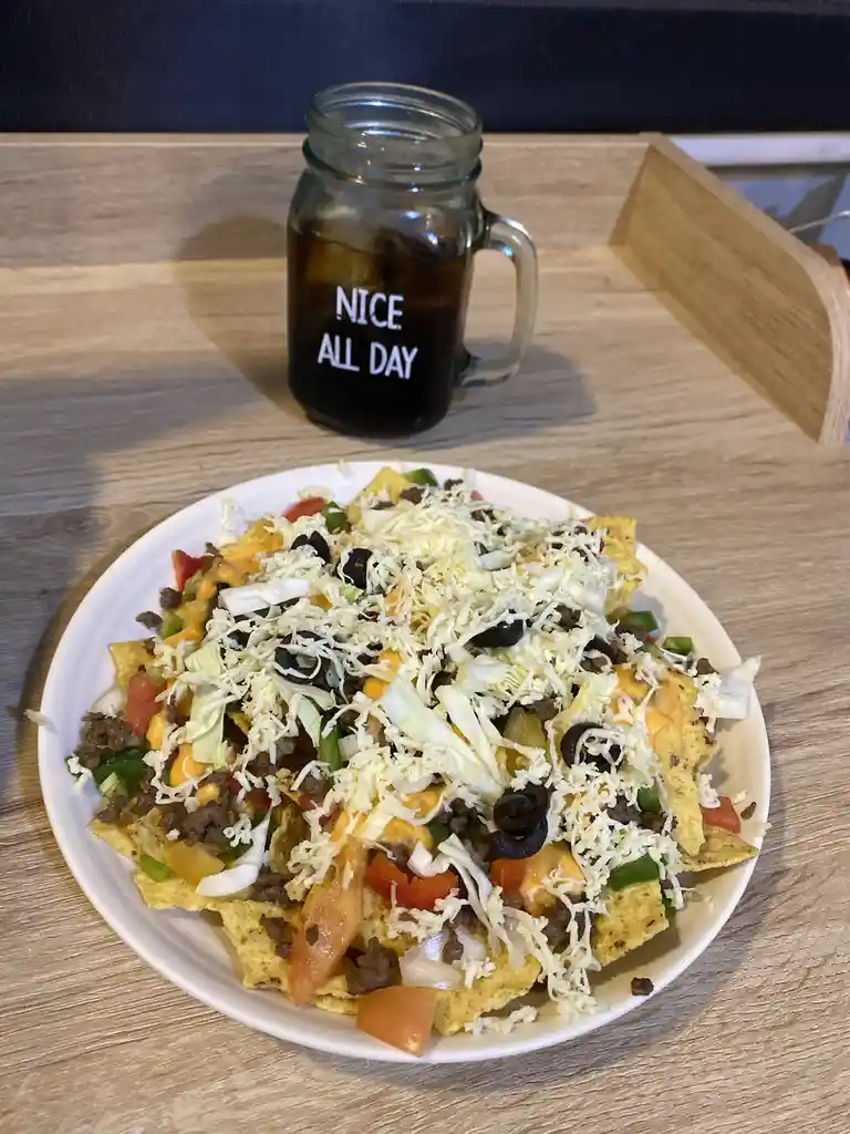 A plate of loaded nachos with cheese, meat, tomatoes, and olives sits on a wooden table next to a mason jar mug filled with…