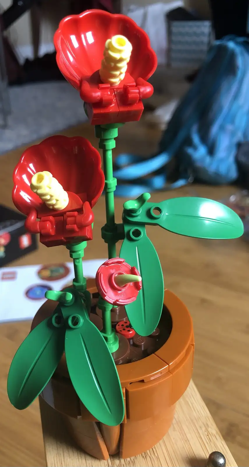 a close-up photo of a lego plant with red flowers, green leaves, and a brown pot, sitting on a wooden surface. A small red…