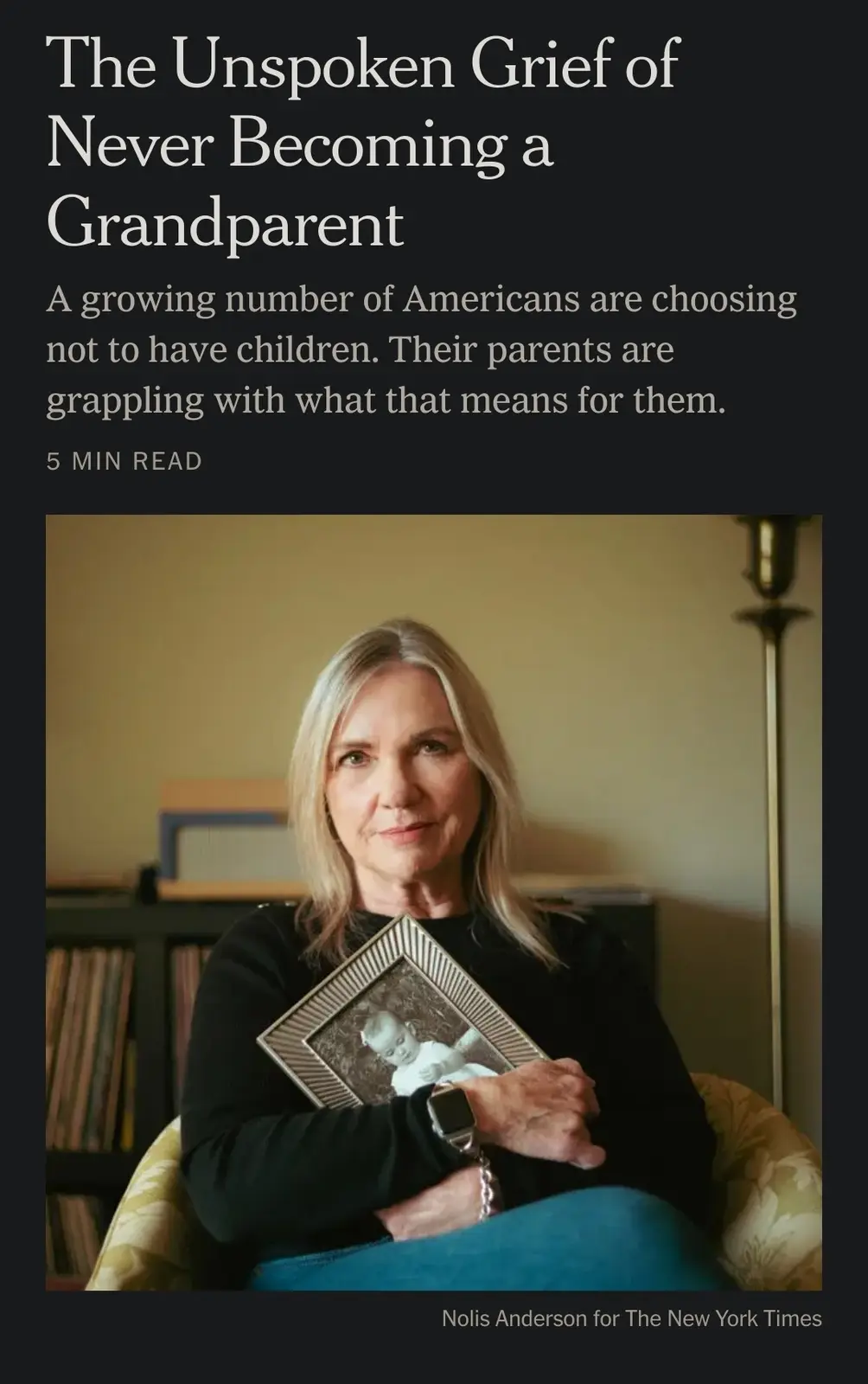 A somber-looking middle-aged woman with blonde hair sits in a chair holding a framed photo of a baby, wearing a black shirt…