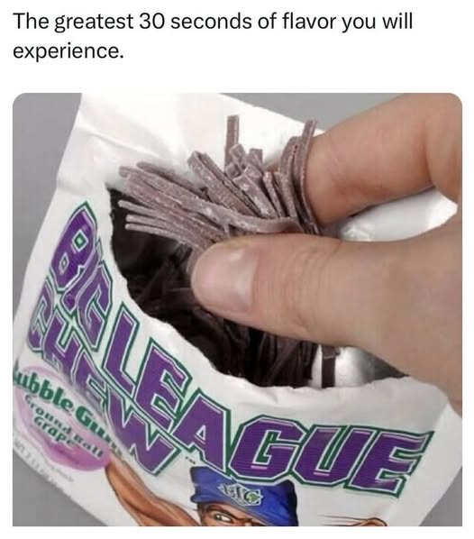 A hand pulls out a handful of purple, stringy, chewy candy from a torn-open bag labeled 'Big League Chew' with 'Grape'…