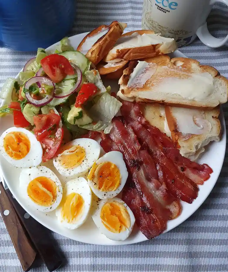 A plate of breakfast food including sliced hard-boiled eggs, crispy bacon, buttered toast, and a fresh salad with tomatoes…