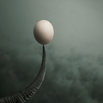 A pale, smooth egg is balanced precariously on the sharp, curved tip of a dark, textured tusk against a muted gray-green…