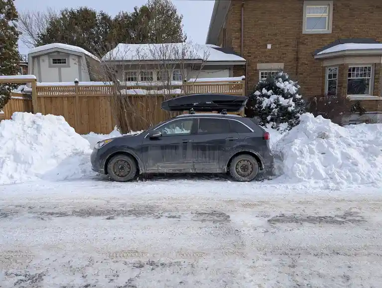 A dark gray SUV is parked in a snowy driveway, flanked by large snow piles on both sides. The car has a black Thule roof box…