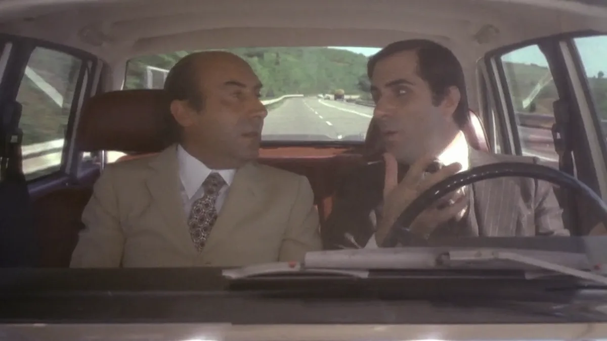 Two men are in a car; the driver, holding a cigarette, looks shocked while the passenger in a beige suit stares ahead. The…