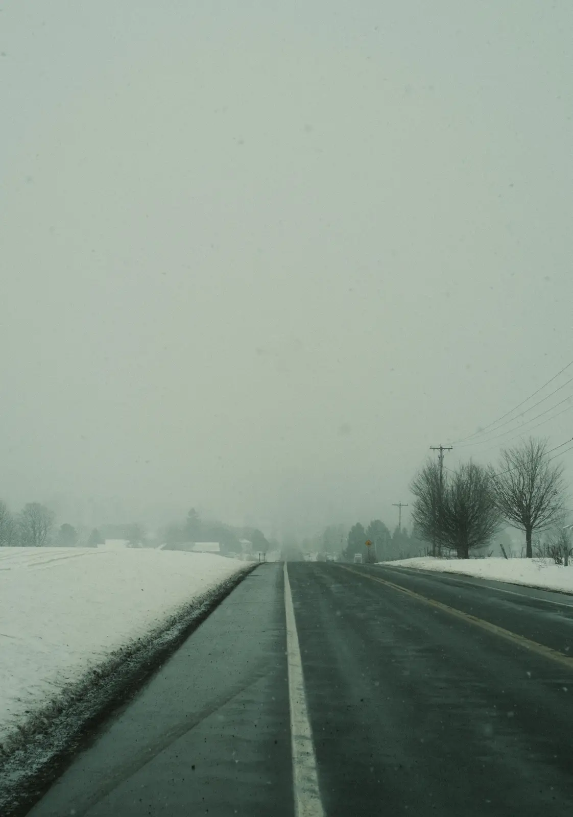 A desaturated, snowy road stretches into a foggy, gray distance with snow-covered fields on either side and bare trees…