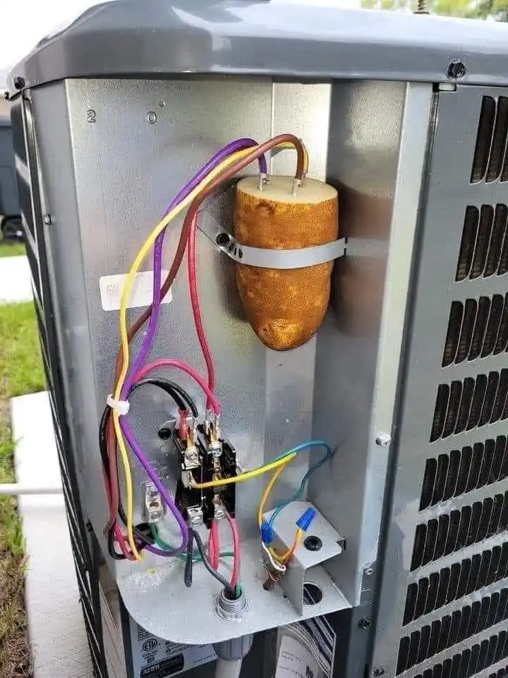 A large, irregularly shaped potato is strapped to the inside of an outdoor HVAC unit, with multiple colorful wires connected…