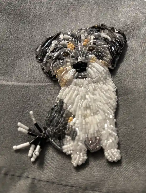 A handmade beadwork art piece depicting a small dog with black, white, and translucent beads, sitting on a gray fabric…