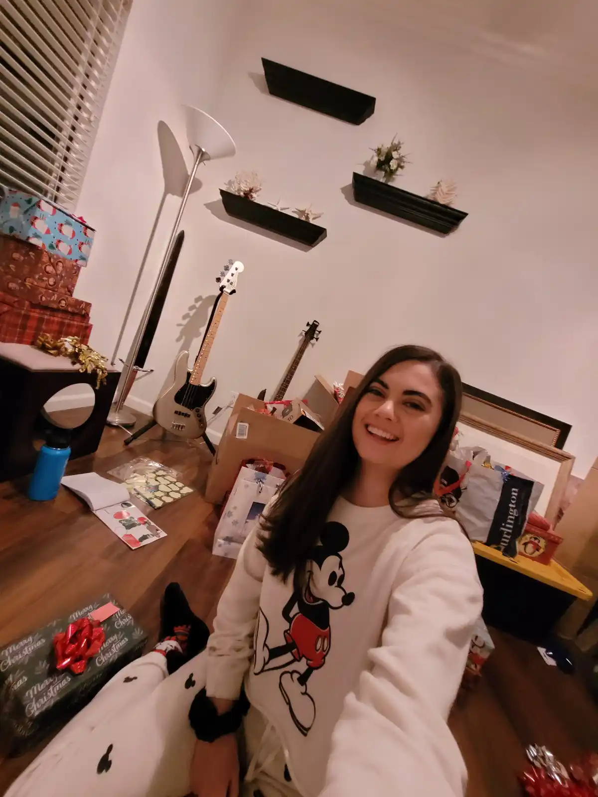 A woman in a Mickey Mouse sweatshirt smiles at the camera while sitting on a wooden floor surrounded by Christmas presents…