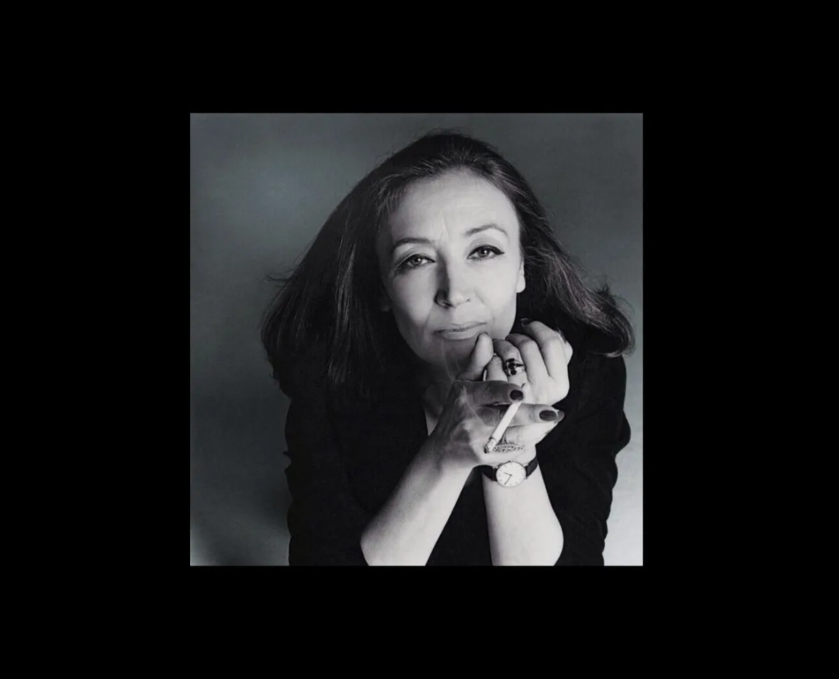 a black and white portrait of a woman with dark hair, wearing a black top, holding a cigarette between her fingers near her…