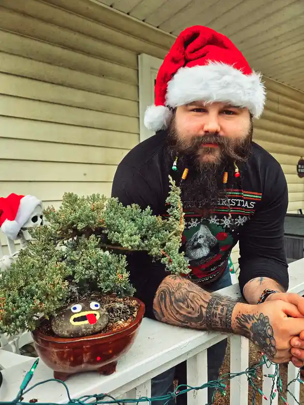 A man with a long beard wearing a red Santa hat and a Christmas sweater leans on a white porch railing next to a bonsai…