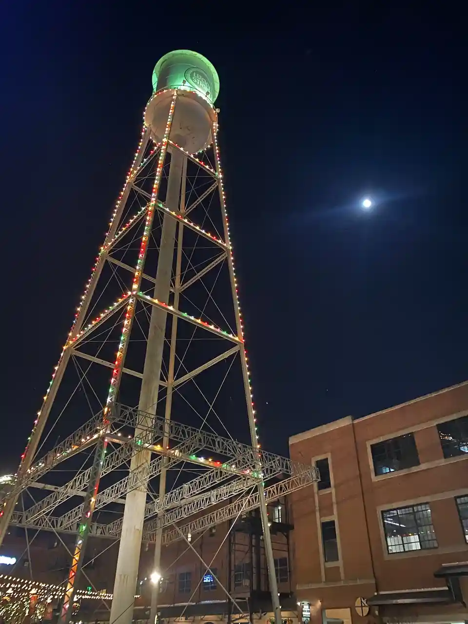 A tall, metal water tower at night, decorated with colorful string lights, has a green-lit top with the words 'LUCKY STRIKE'…