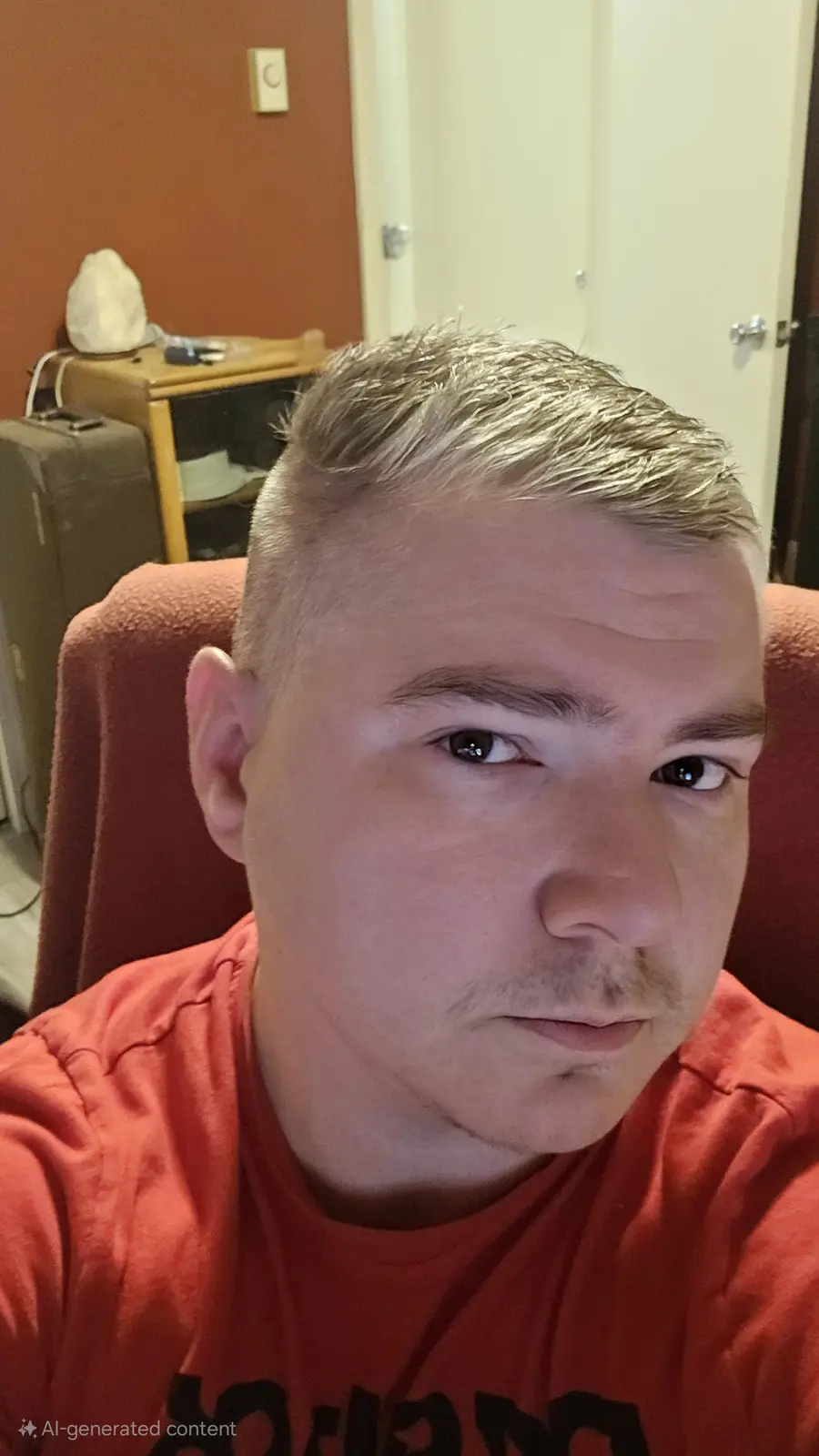 A close-up selfie of a man with a faded buzz cut, wearing an orange t-shirt, sitting in a red chair indoors. The background…