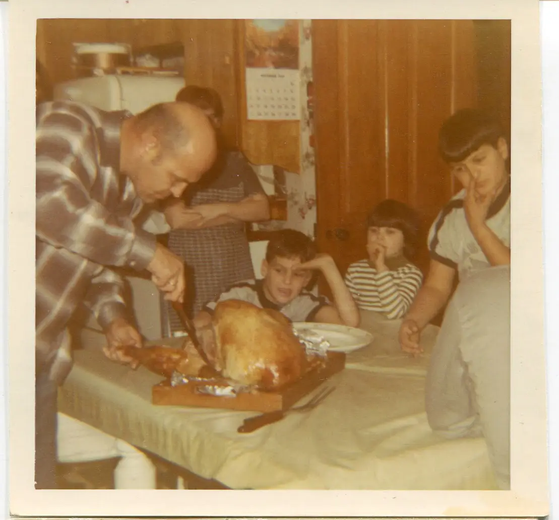 A vintage photo shows a man carving a roasted turkey on a wooden board at a dining table, surrounded by several children…