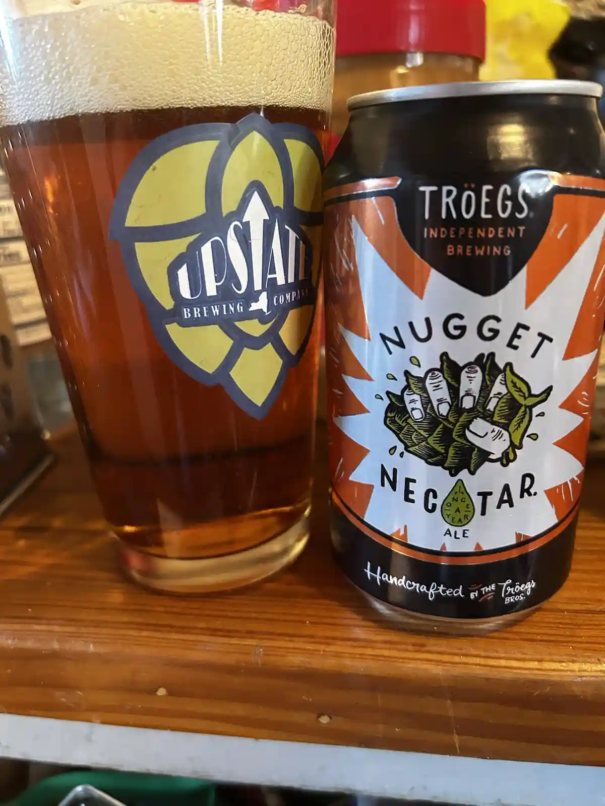 A pint glass with an 'Upstate Brewing Company' logo sits next to a can of 'Troegs Independent Brewing Nugget Nectar Ale' on…