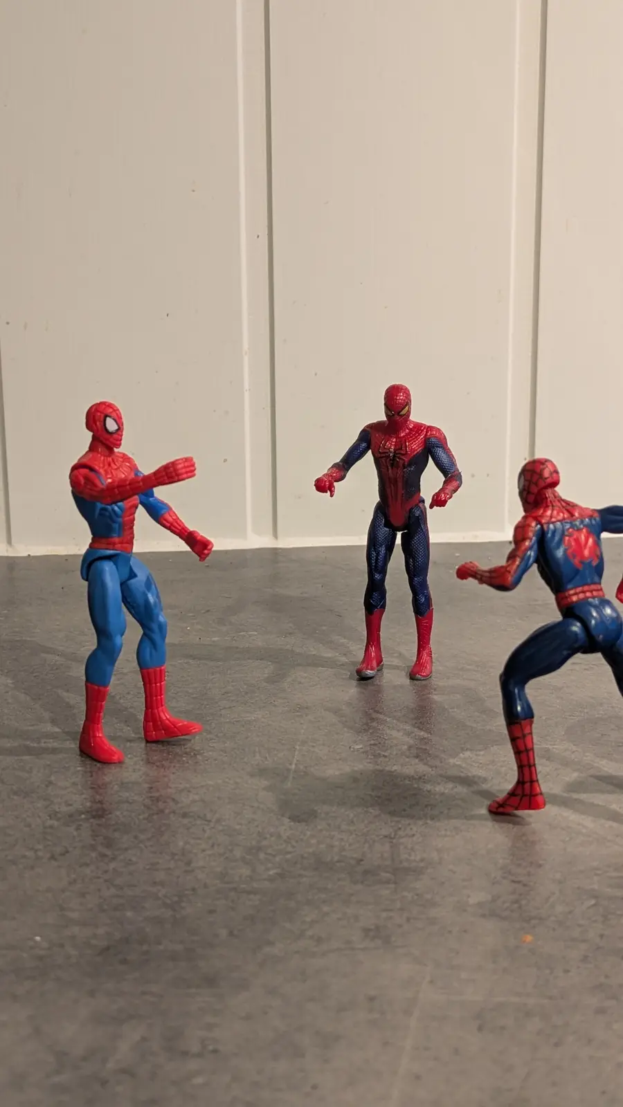 Three Spider-Man action figures stand on a gray floor against a white paneled wall. The central figure, in a darker, more…