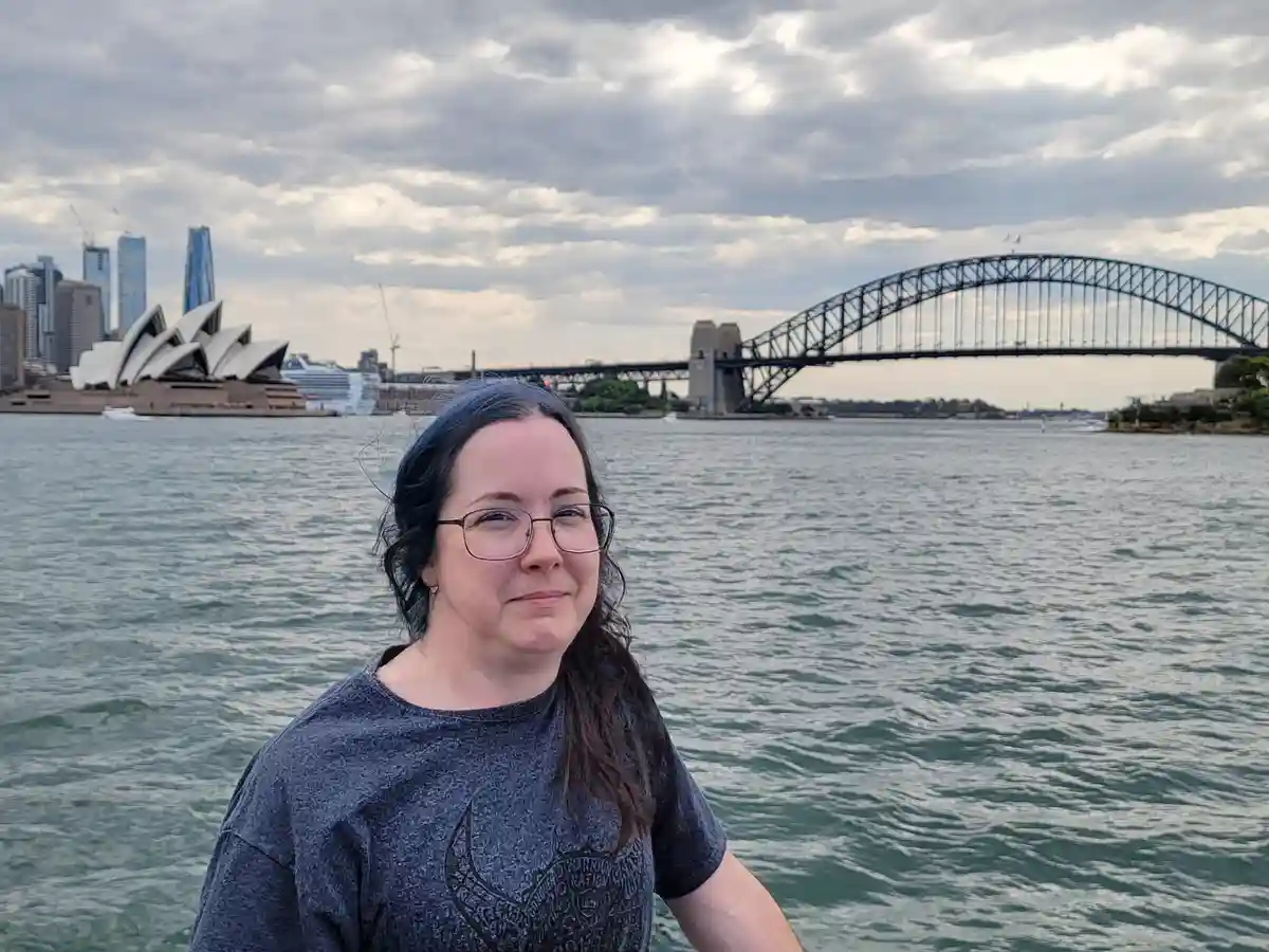 A person with glasses and dark hair stands in front of a body of water, with the Sydney Opera House and Sydney Harbour…